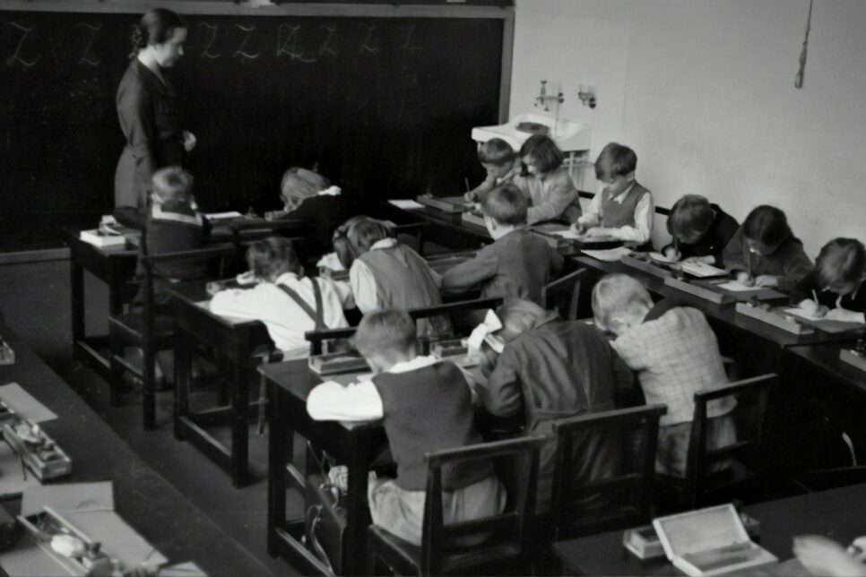 grayscale photography of teacher standing near chalkboard and children sitting on chairs